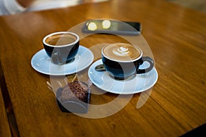 two cups of cappuccino on black cups, on wooden table, with typical brasilian sweet called brigadeiro. Cell phone on the backgroun photo
