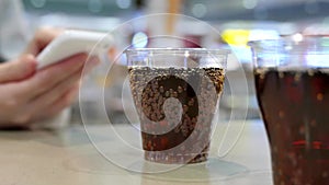 Two cups of coca cola on table with woman reading message on iphone at food court