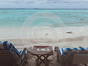 Two cups of coffee on a wooden table and two empty sunbeds. Maldives, the white sand beach near the turquoise ocean.