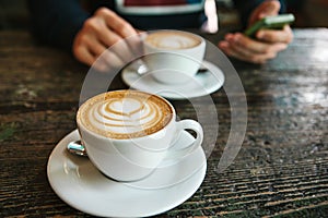 Two cups of coffee on a wooden table, a man holding a phone in his hand and going to call. Waiting for a meeting