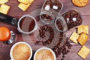 Two cups of espresso, crackers, cookies, holder with ground coffee, tamper and cans of coffee beans on wooden table