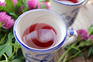 Two cups of healing herbal tea or infusion with red clover, purple flowers on the wooden table. Natural floral background.