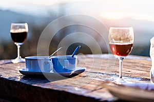 Two cups of hot coffee and two glass of red wine are on the old wooden table which is outdoor among of sunlight in the morning for