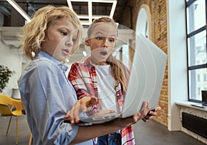 Two curious kids, little boy and girl looking surprised while using laptop, standing in a classroom during STEM lesson