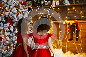 Two curly little girls looking at the christmas fireplace near beautiful Christmas tree. Twins in red dresses looking at