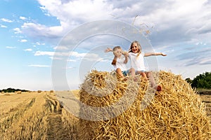 Two cute adorable caucasian siblings enjoy having fun sitting on top over golden hay bale on wheat harvested field near