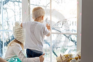 Two cute adorable little blond cauasian children siblings stay near window and looking outside waiting for snow, wonders photo