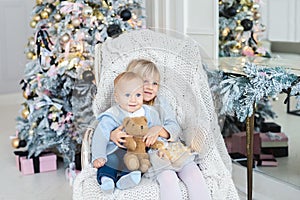 Two cute adorable little siblings brother and sister sitting in chair near christmas tree in cozy living room looking , dreaming