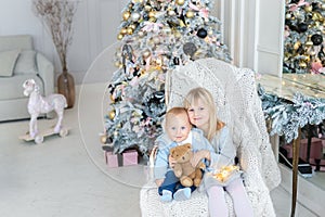 Two cute adorable little siblings brother and sister sitting in chair near christmas tree in cozy living room looking , dreaming