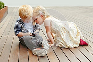 Two cute adorable white Caucasian toddlers boy and girl sitting together and playing games on cell mobile phone digital tablet.