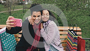 Two cute african american woman taking selfie on smartphone with shopping bags and smiling. Girlfriends sitting on