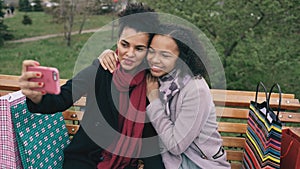 Two cute african american woman taking selfie on smartphone with shopping bags and smiling. Girlfriends sitting on