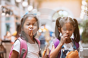 Two cute asian child girls with backpack eating pancake together after school