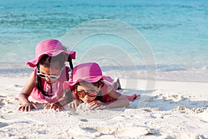 Two cute asian child girls wearing pink hat and sunglasses playing with sand together on the beach near the beautiful sea