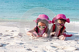 Two cute asian child girls wearing pink hat and sunglasses playing with sand together on the beach near the beautiful sea