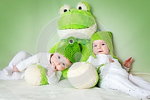 Two cute babies lying in frog hats with a soft toy