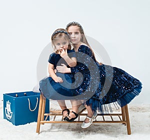 two cute baby girls sisters with blue eyes wear dark blue dresses sit on a decorative bench and hug