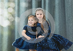 two cute baby girls sisters with blue eyes wear dark blue dresses sit on a decorative bench and hug