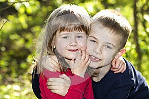 Two cute blond funny happy smiling children siblings, young boy brother embracing sister girl outdoors on bright sunny green bokeh