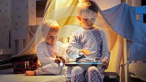 Two cute boys in pajamas playing games on tablet computer in bed at night. Children with gadgets, education, kids development