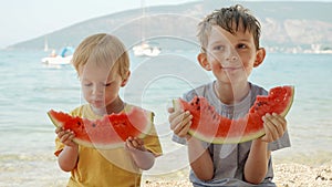 Two cute boys sharing sweet watermelon on a beach towel. Summertime fun, holiday happiness, and vacation relaxation