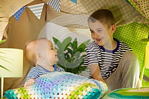 Two cute brothers play with a flashlight in a children`s tent. 6 years old boy and baby toddler