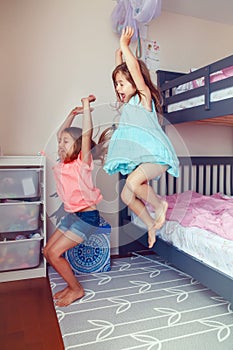 Two cute Caucasian girls siblings jumping from bed in room. Happy excited friends having fun at home. Adorable children playing