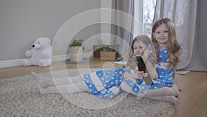 Two cute caucasian girls sitting on soft carpet indoors with remote control. Twin sisters in blue dotted dresses