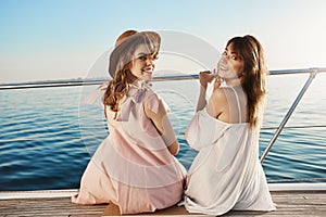 Two cute european female friends sitting at side of boat, turning back to look at camera while smiling broadly in good