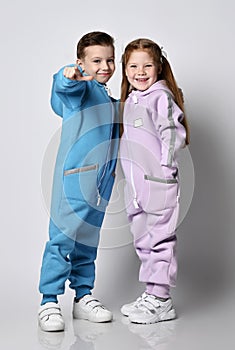 Two cute, frolic kids in jumpsuits are standing face to face and boy is pointing with finger at camera