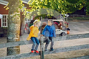 Two cute funny Caucasian children boy girl sitting on wooden rustic fence in park farm outside