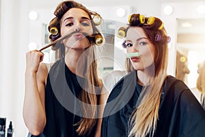 Two cute girlfriends in black cape wearing curlers making moustache out of their hair and roller in hairdressing salon