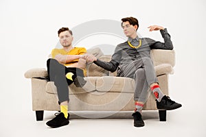 Two cute guys make a bet hand in hand sitting on a white sofa. Group of friends are sitting on a soft couch and