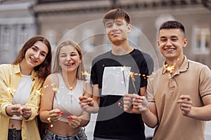 Two cute guys and two charming girls are holding sparklers and smiling. Friends in casual clothes have fun and laugh