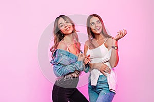 Two cute happy women with positive smiles in trendy youth jeans clothes posing and smiling on background glamorous pink wall in