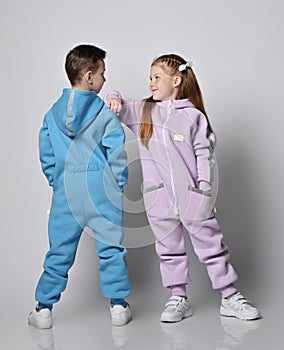 Two cute kids in blue and pink jumpsuits are standing next to each other, boy with his back to us and girl with her face