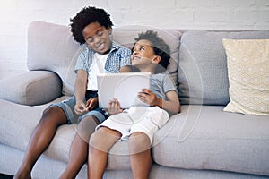 Two cute little african american boys sitting comfortably on a sofa using a wireless tablet while browsing and smiling