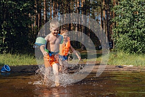 Two cute little boys running into lake splashing water in the forest. one boy wearing life vest, amother wearing armbands.