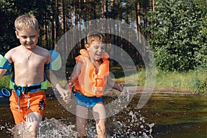 Two cute little boys running into lake splashing water in the forest. one boy wearing life vest, amother wearing armbands.