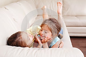 Two cute little Caucasian girls siblings playing at home. Adorable smiling children kids lying on a couch together. Authentic