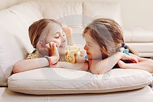 Two cute little Caucasian girls siblings playing at home. Adorable smiling children kids lying on a couch together. Authentic