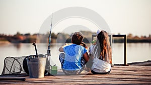 Two cute little friends, boy and girl eating sandwiches and fishing on a lake in a sunny summer day