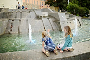 Two cute little girls playing by city fountain on hot and sunny summer day. Children having fun with water in summer