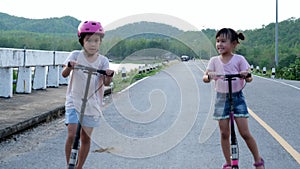 Two cute little girls riding kick scooters on outdoor road in summer park.