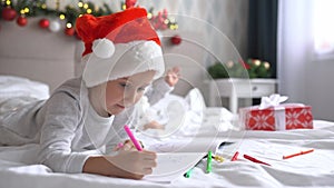 Two cute little girls sisters wearing Santa Helper hats write letter to Santa lying on bed at home in Christmas