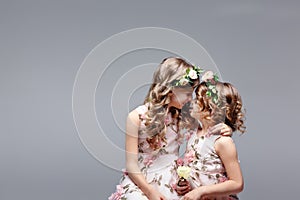 Two cute little girls - sisters in wreaths of fresh flowers on their heads sit and hug in a bright studio. Family and childhood