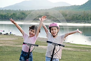 Two cute little girls smiling and posing together in summer garden. Happy kid riding kick scooter in the park. Healthy sports and