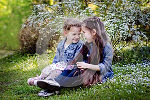 Two cute little girls tell some secrets each over while sitting on the grass in a spring park
