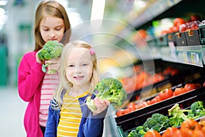 Two cute little sisters choosing broccoli in a food store or a supermarket