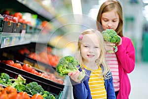 Two cute little sisters choosing broccoli in a food store or a supermarket